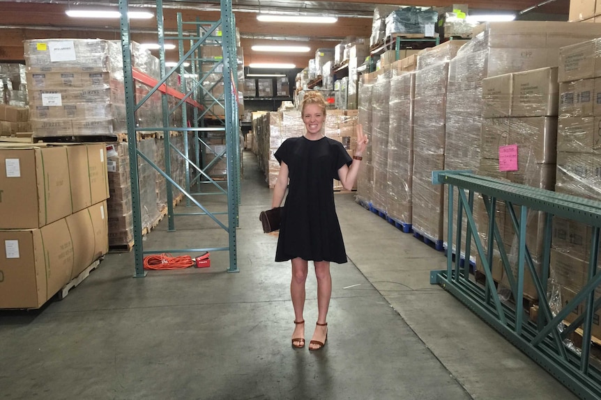 A woman smiles broadly and gives a peace sign, surrounded by cardboard boxes in a warehouse