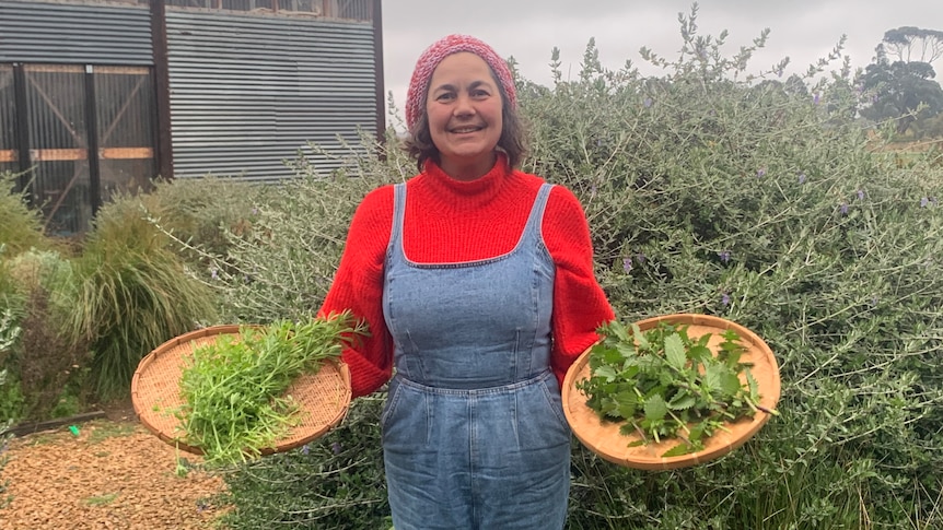 A woman in a garden holds two baskets full of green weeds, she is smiling at the camera