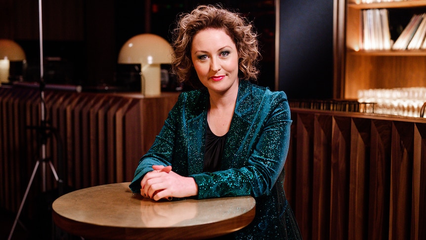 Zan Rowe sits at a table in a dim room, wearing a sparkly green blazer, looking at the camera, with her hands clasped