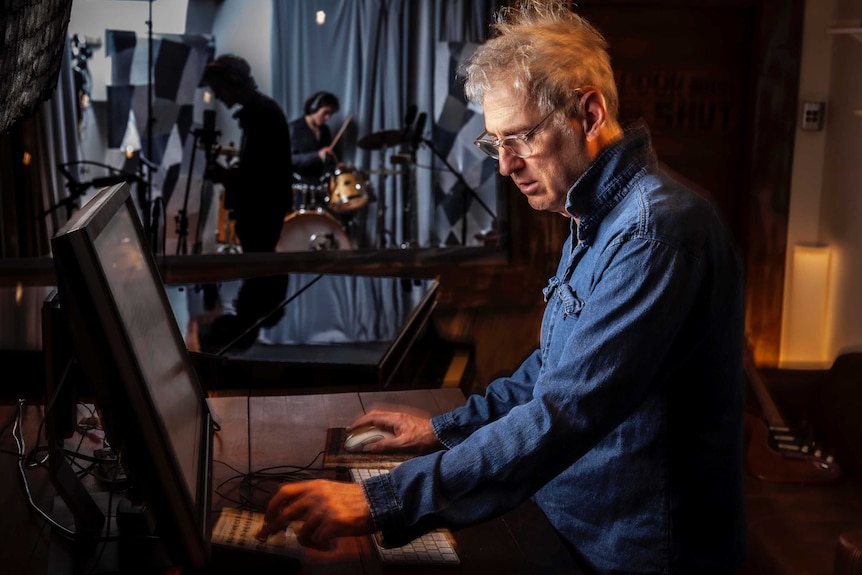 A man sits at a panel desk with musicians in the background.