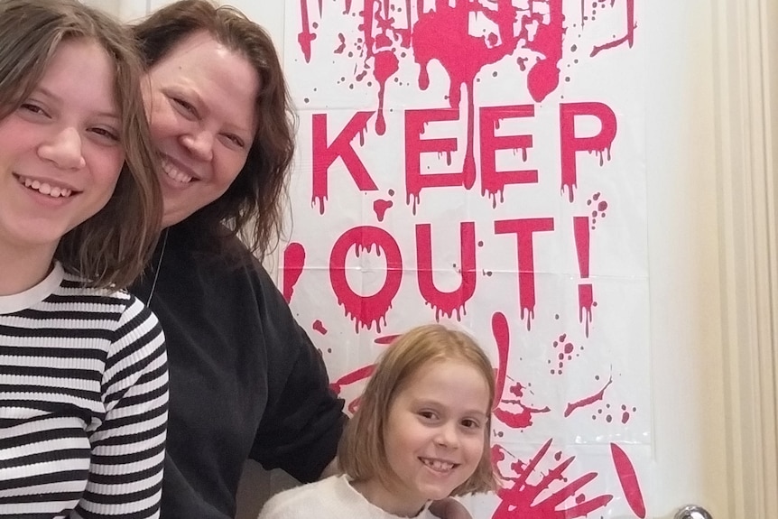 A woman and two children stand in front of a door with a Halloween-themed sign saying 'keep out'.