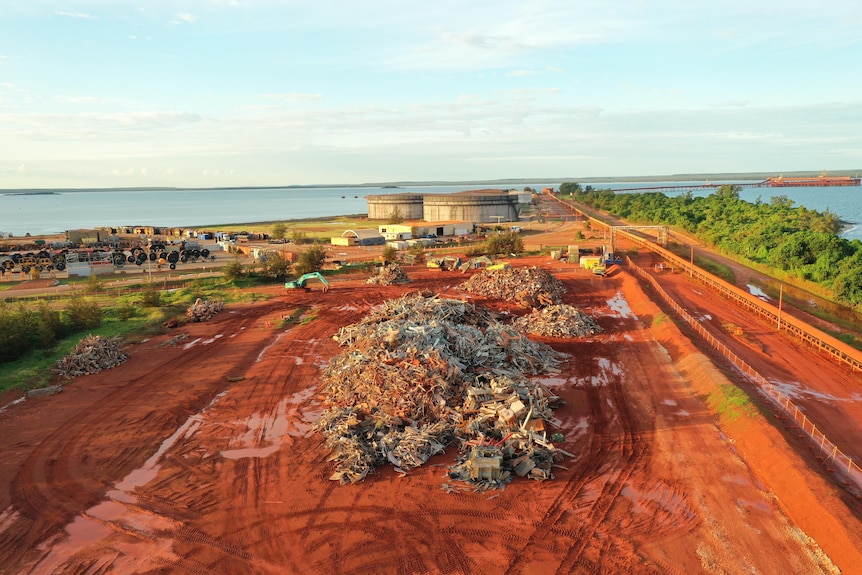a pile of scrap metal on red dirt with tanks and a harbour in the background.