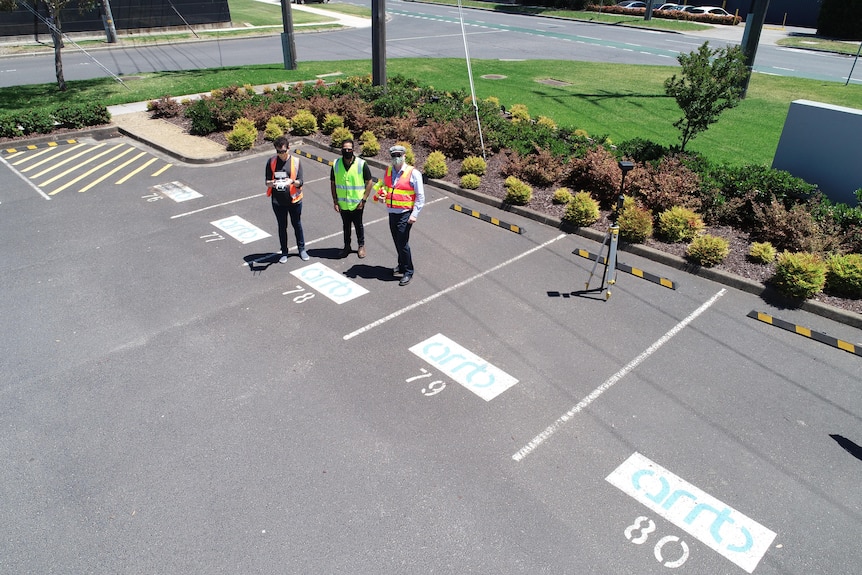 Three men stand in a car park, photographed by a drone in the air.