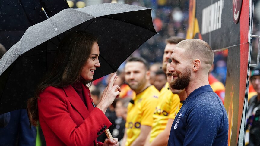 一位公主微笑着交叉手指，她在比赛前的雨中与一位英国橄榄球联盟球员交谈。 