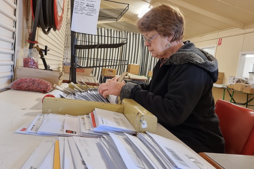 Woman in black jacket sitting down at a desk sorting piles of envelopes.