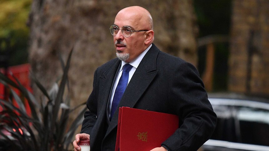 A bald man with a dark suit holds a folder.