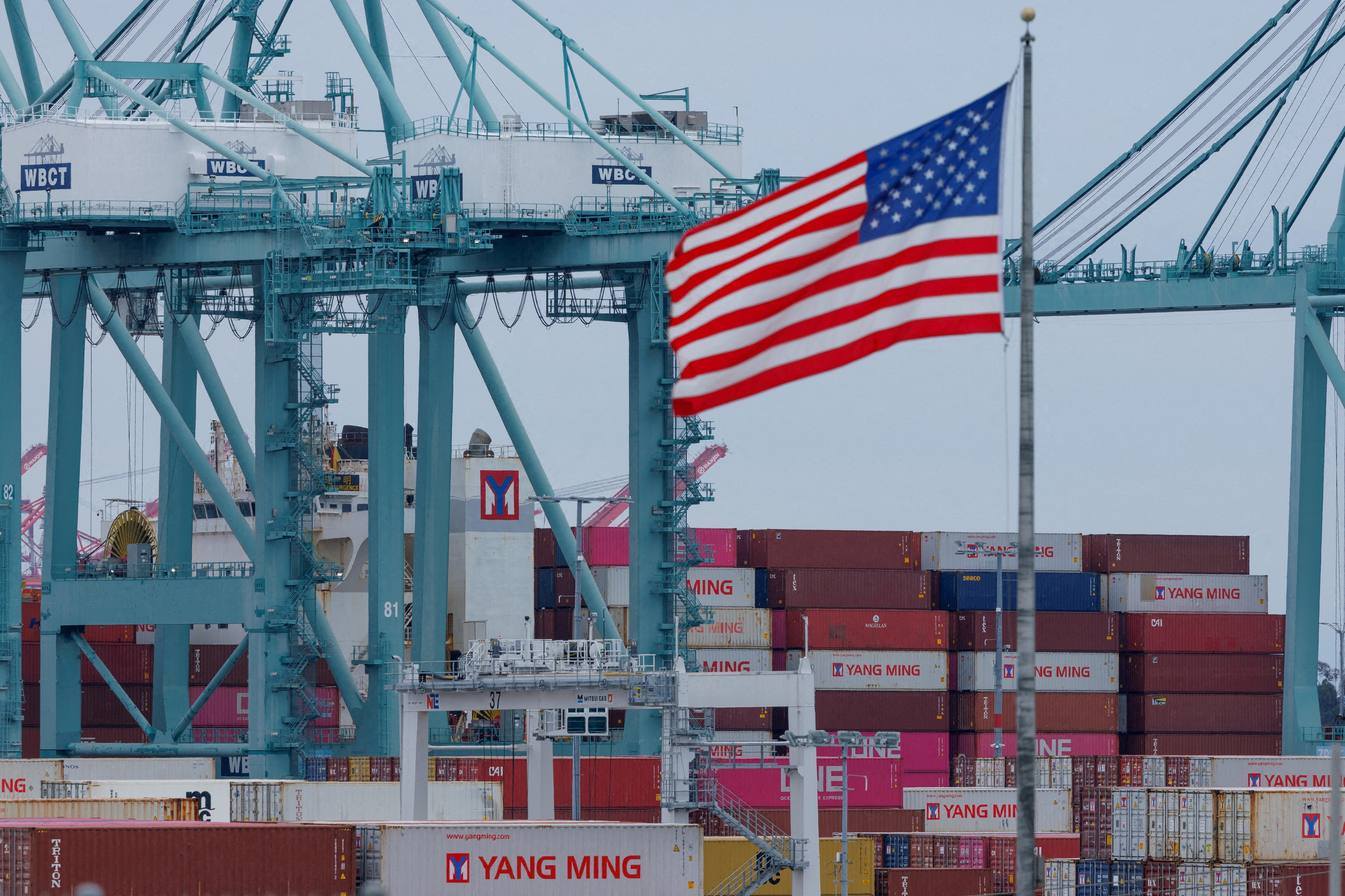 An American flag flying in front of stacks of shipping containers at a port.