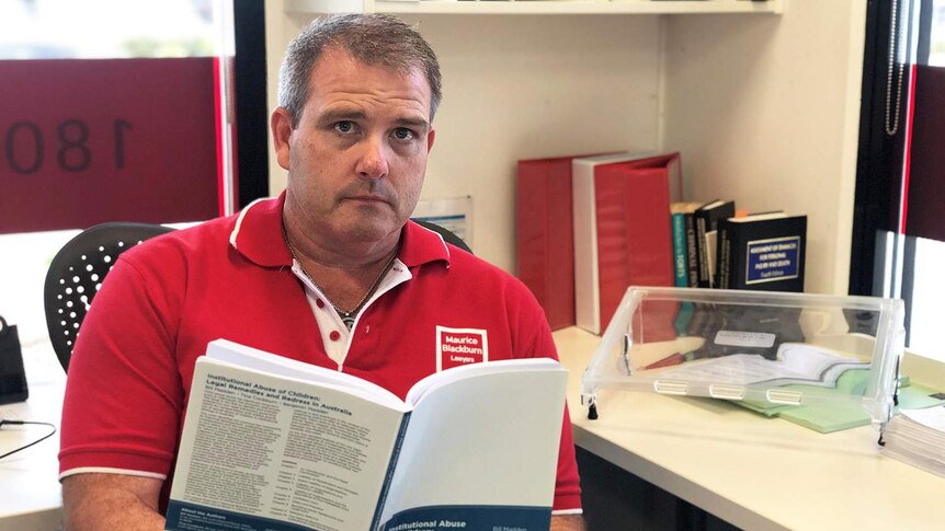 Lawyer Jed McNamara looks at the camera as he sits at a desk holding a report at his Maroochydore office.