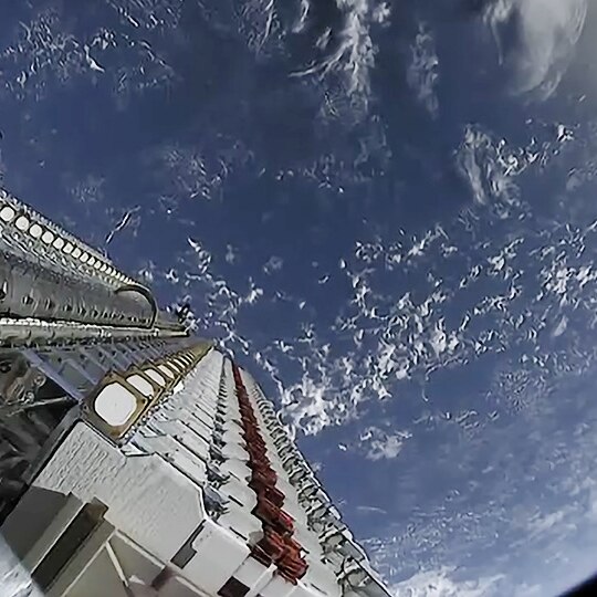 A camera on board a satellite looks down towards Earth showing part of the satellite and the planet.