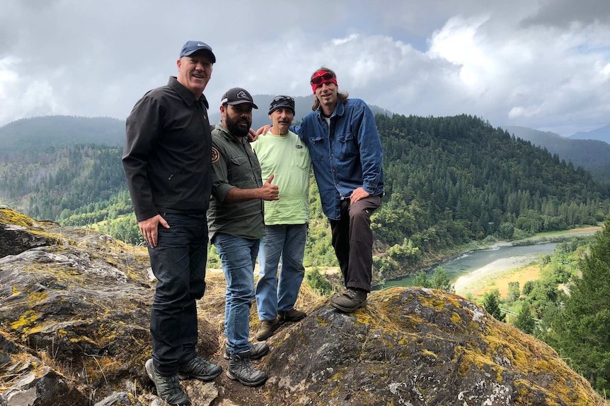 Scott Falconer stands with three men on top of a hill over looking a river and forest on a cloudy day