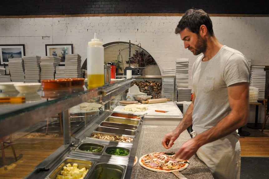 Tim Boyle stands behind a counter and puts toppings on a pizza base.