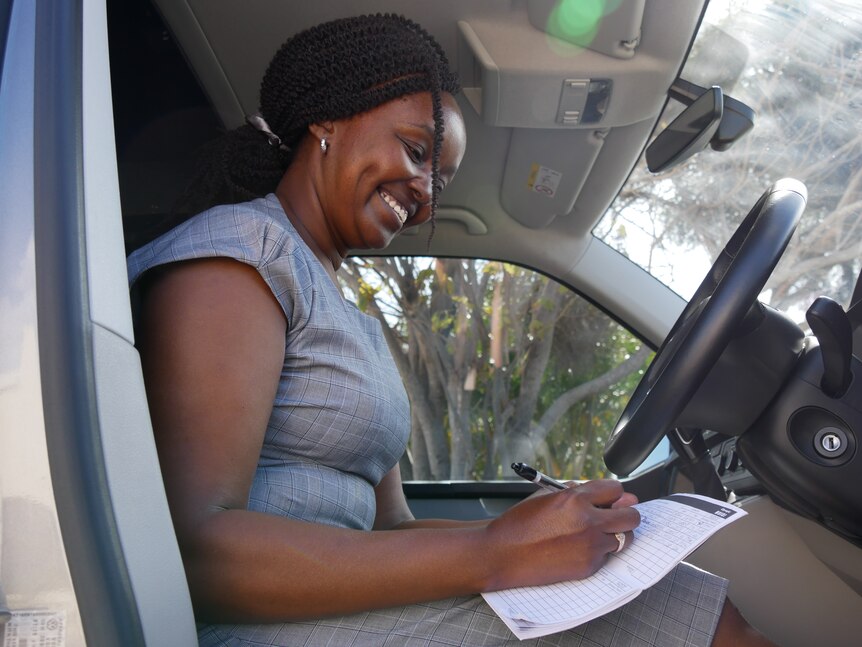 A woman of Kenyan heritage looks down at a car logbook in the drivers seat smiling. 