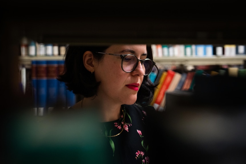 A woman reads a book in an aisle of bookshelves lined with different coloured books in a cluttered library.
