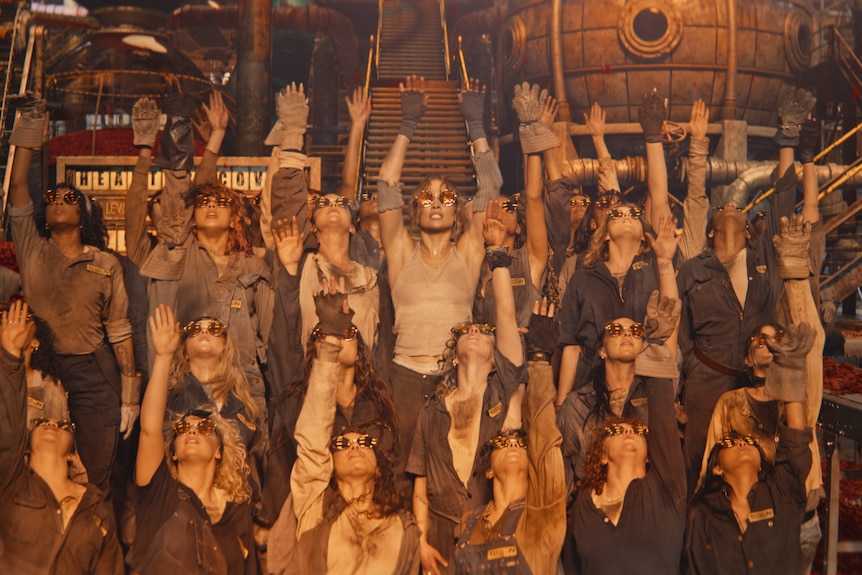 Jennifer Lopez stands among a dancer's chorus in cyberpunk uniforms and goggles inside a factory.