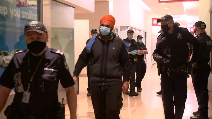 A man wearing an orange turban and a face mask is surrounded by police in an arcade