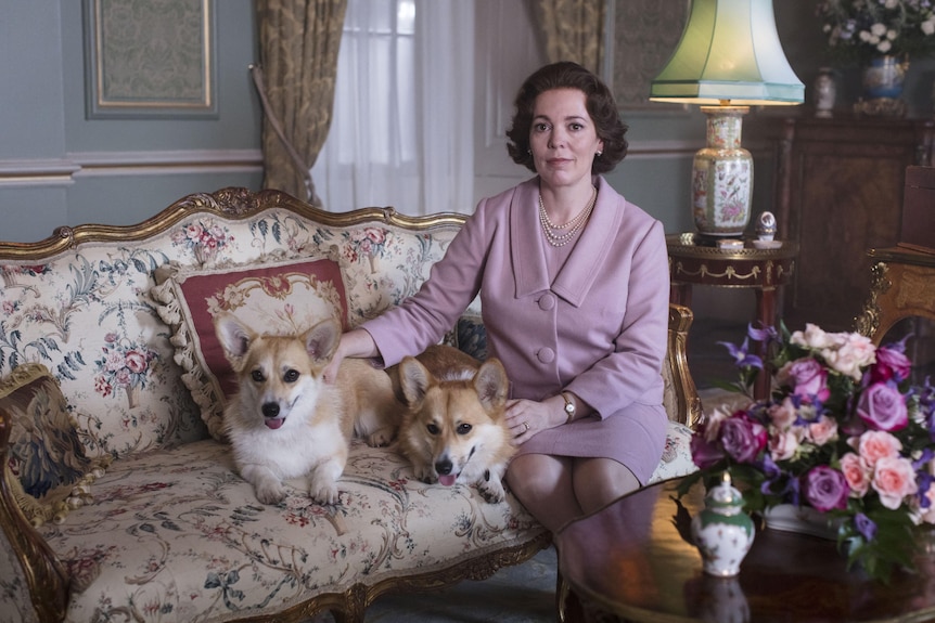 Olivia Colman, dressed as the Queen in a pink outfit, sits in a grand living room on a couch with two smiling corgis