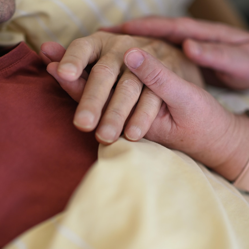 A close up photograph of a hospice worker holding the hand of a terminally ill patient. 