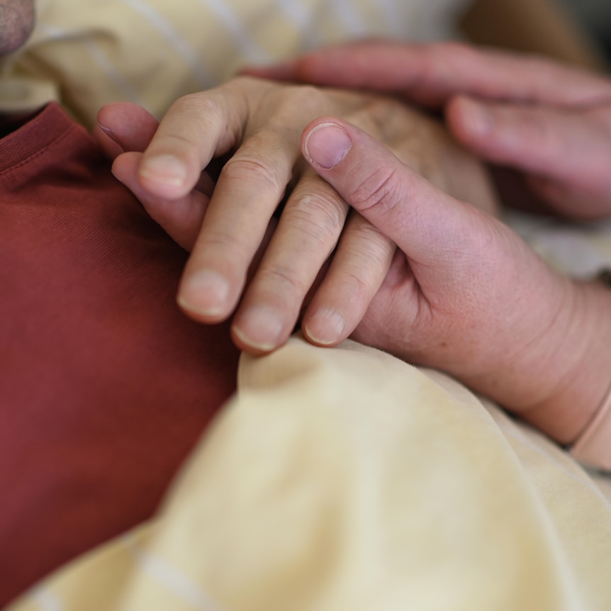 A close up photograph of a hospice worker holding the hand of a terminally ill patient. 