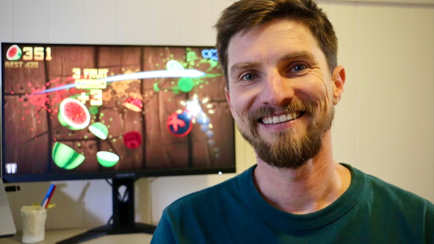 Luke Muscat smiling in front of a screen with a still from Fruit Ninja. Fruit being sliced. 
