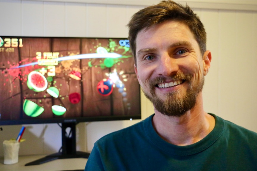 Luke Muscat smiling in front of a screen with a still from Fruit Ninja. Fruit being sliced. 