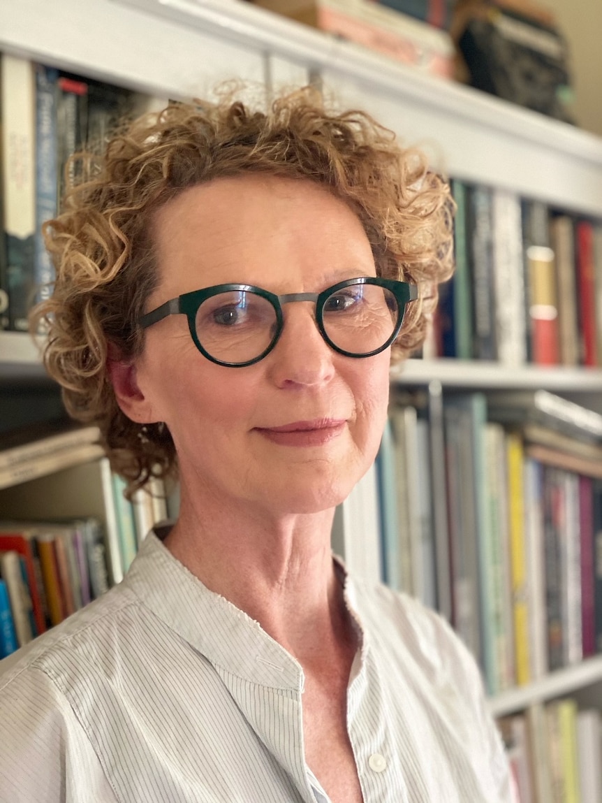 A woman with short, blonde curly hair stands in front of a bookcase.