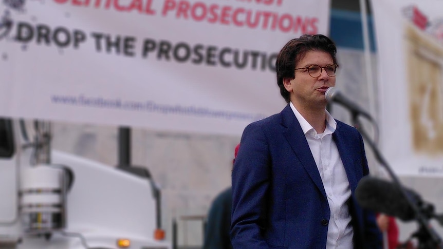 A man addresses a rally in front of a banner saying "Alliance against political prosecutions" 