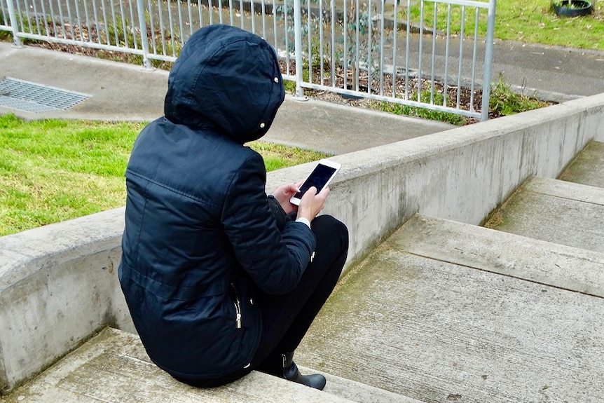Unidentified person sits on a step wearing a face mask and holding a phone.