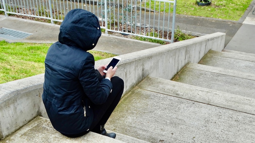 Unidentified person sits on a step wearing a face mask and holding a phone.