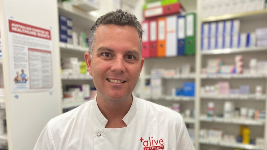 A man wearing a pharmacist's white coat standing in a chemist shop with shelves full of medicine.