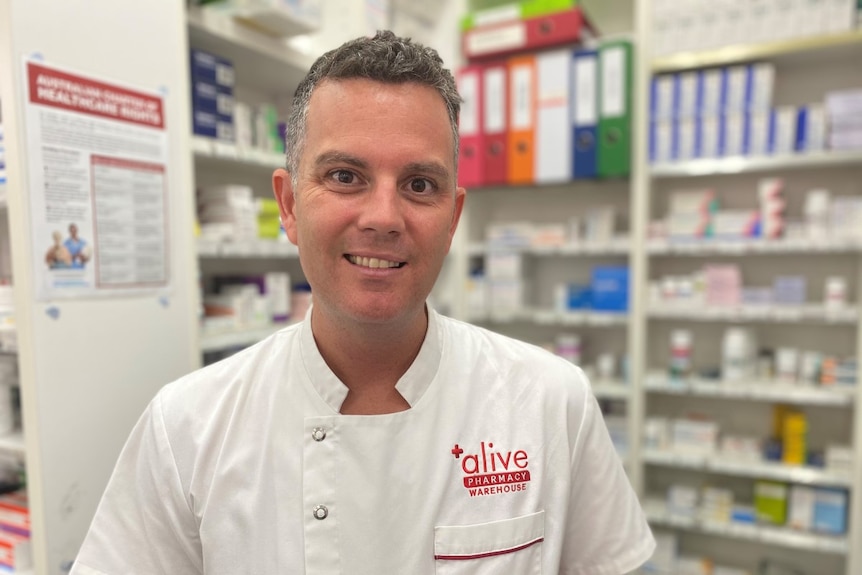 A man wearing a pharmacist's white coat standing in a chemist shop with shelves full of medicine.