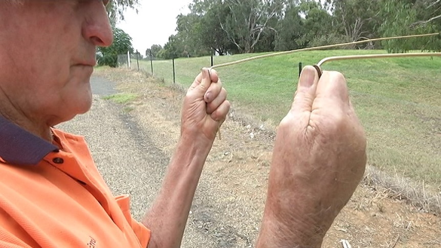 Man holding up two metal rods in his hands, with coins between his two thumbs and the metal rods