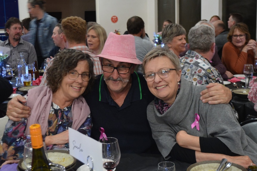 An older man, wearing glasses, grey moustache pink hat, holds two women close, all smile at the camera, wear pink ribbon.