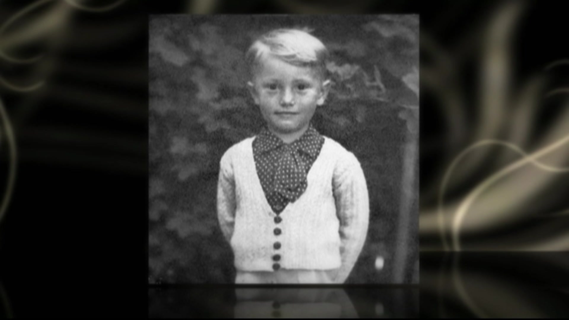 A black and white photo of a light haired boy in a white cardigan over a polka dot collared shirt.