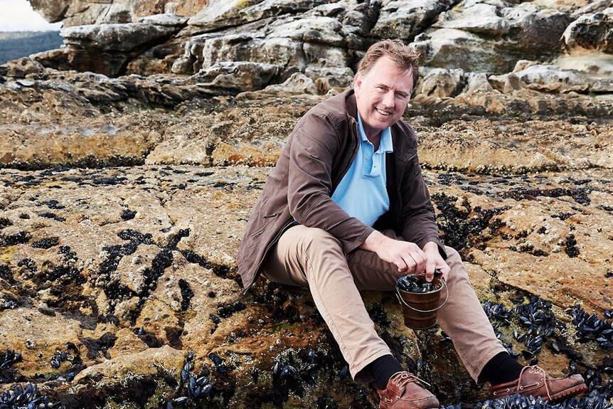 A middle-aged man sits on a rocky shore