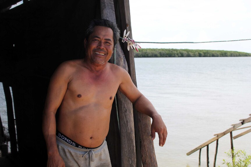 a man without a shirt standing next to his shack near a river.