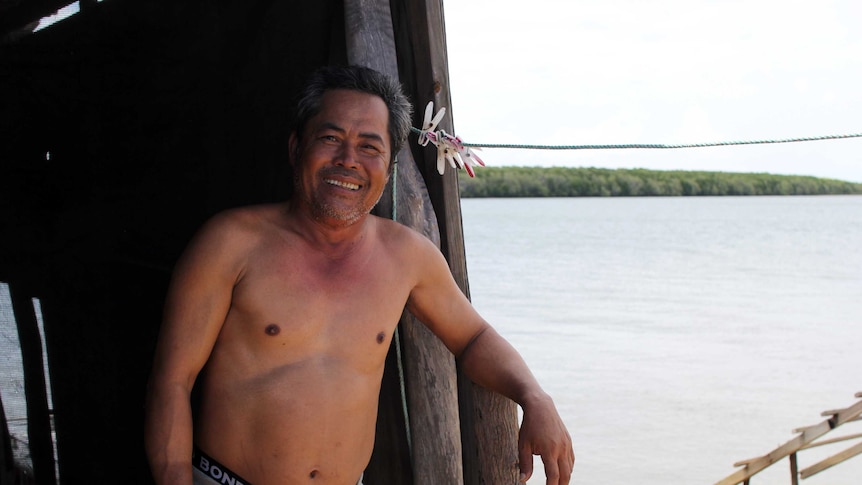 a man without a shirt standing next to his shack near a river.