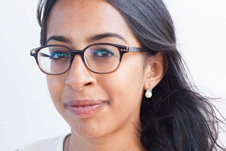 A woman with long dark hair, brown eyes, glasses, pearl earrings stares directly into the camera.