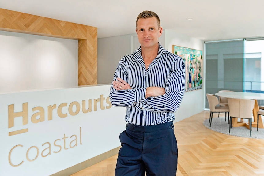 A man in a striped shirt stands cross-armed in an office in front of a Harcourts Coastal sign