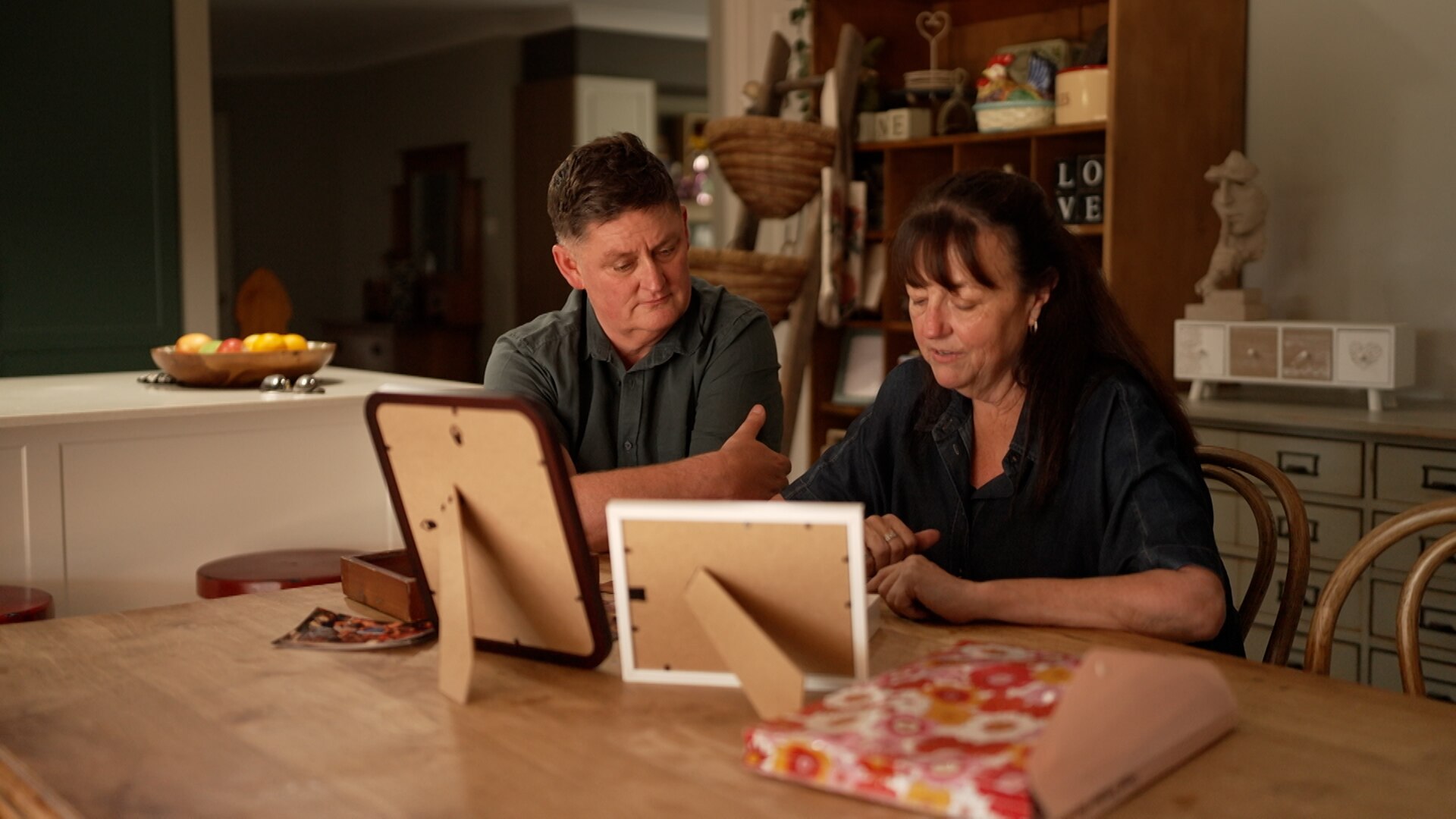 A man and a woman seated at a kitchen table look at framed and album photographs.