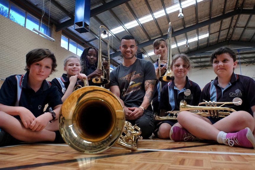 A ground-level photo of singer Guy Sebastian and 6 school children sitting on a gym floor with trumpets and trombones.