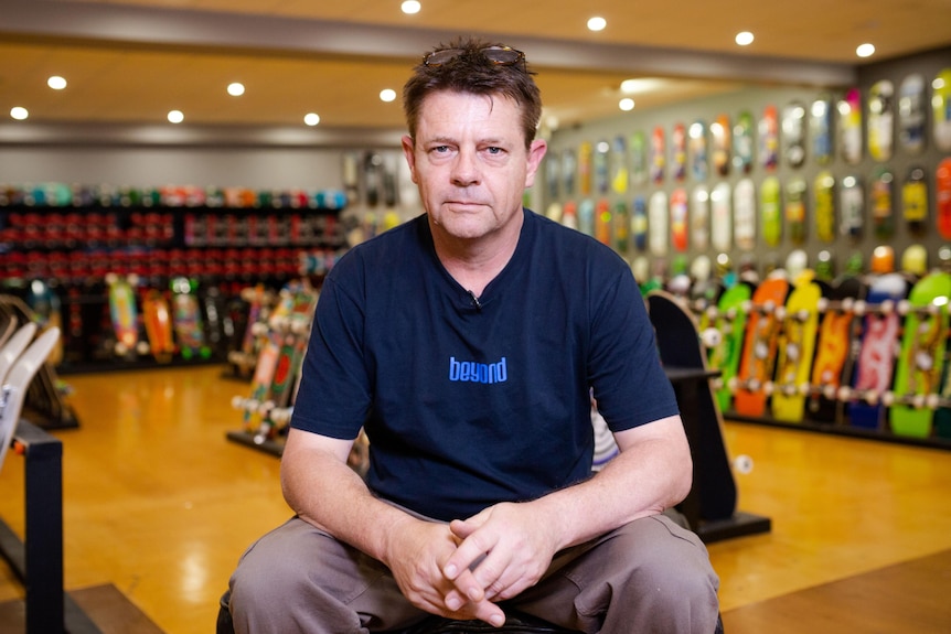 A man stares down the barrel of the camera. Skateboards in all their colourful glory line the walls behind.