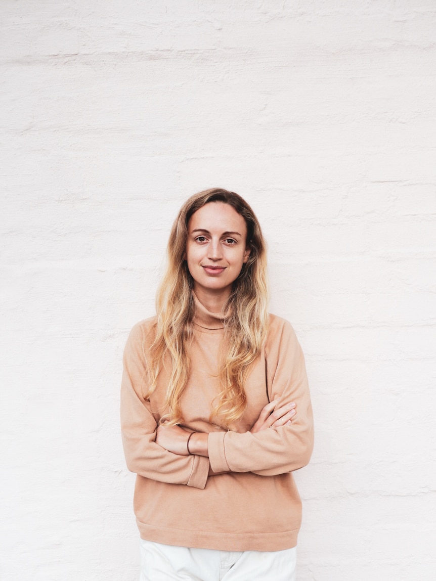 A white woman in her early 30s with blonde hair wearing a light pink skivvy and standing in front of a white wall