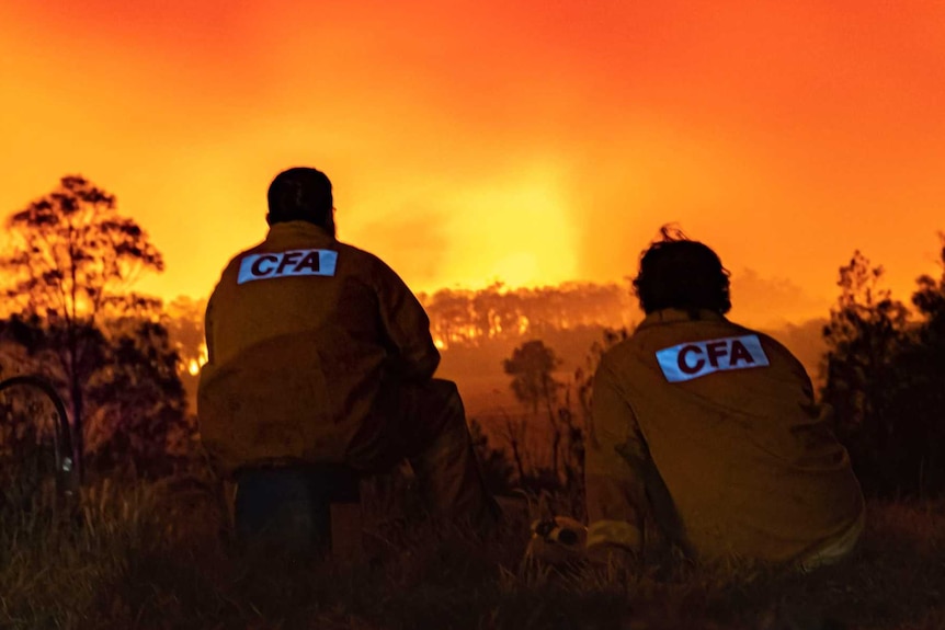 Two CFA volunteers with their backs to the camera, sit crouched on the ground watching a fire burn. The sky is red.