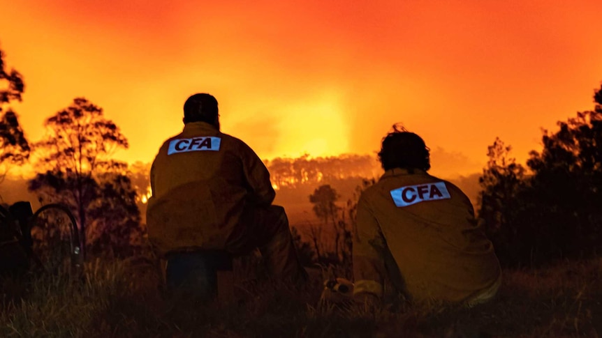 Two CFA volunteers with their backs to the camera, sit crouched on the ground watching a fire burn. The sky is red.