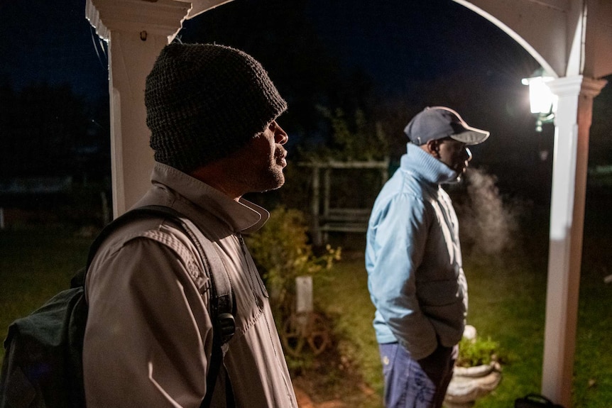 Two men stand on a porch with jackets and hats on, a light is on and the sky is dark. You can see one man's breath in the cold.