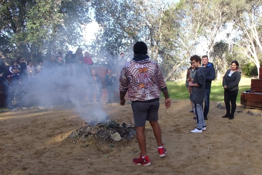 An Aboriginal man in a NAIDOC week jacket attends a smoking fire pit as a group of people watch from seats nearby