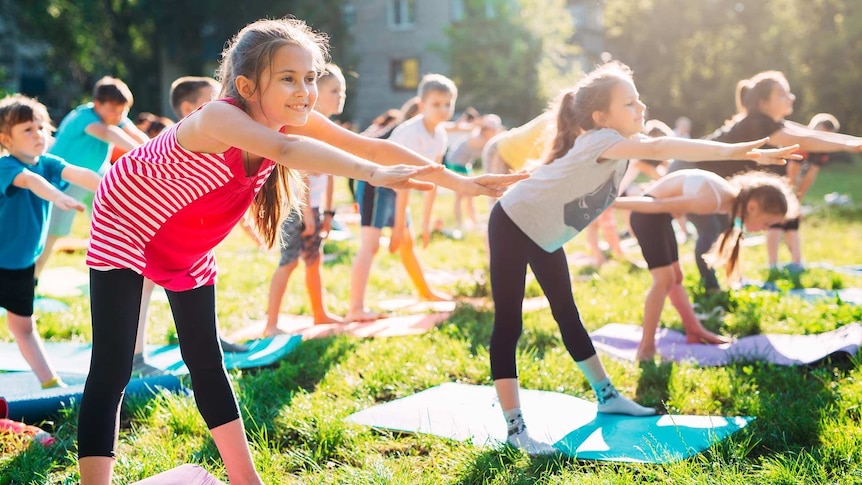 A class of primary school children stand on yoga mats outdoors on grass on a bright sunny day, and they are stretching forwards.