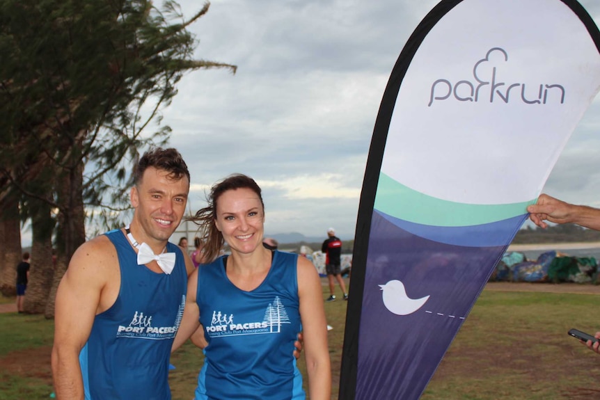 A man and woman in branded parkrun running gear stand smiling together next to a parkrun flag in a park.
