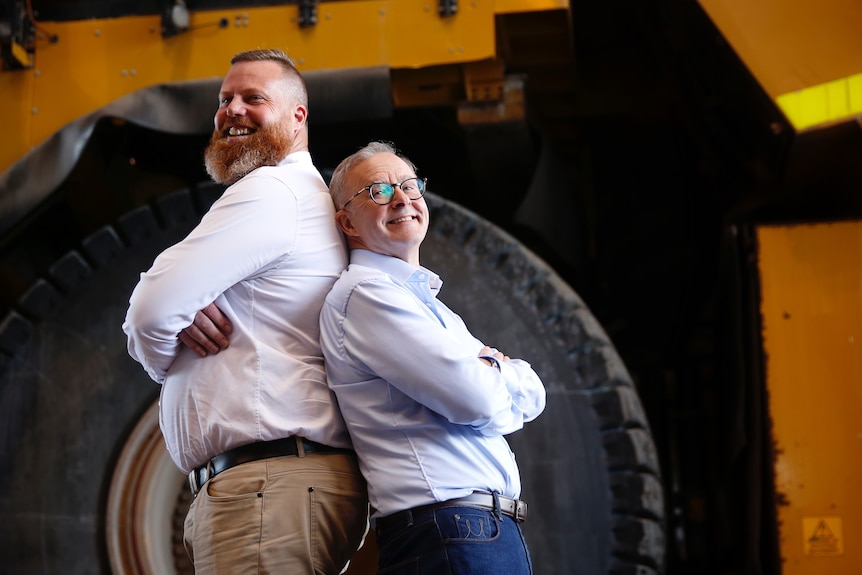 Anthony Albanese stands back to back with a very tall man in front of a big wheel.