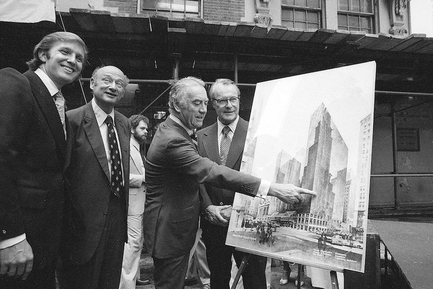 A black-and-white photo shows young Donald Trump smiling next to other men in suits, one pointing at a picture of a skyscraper